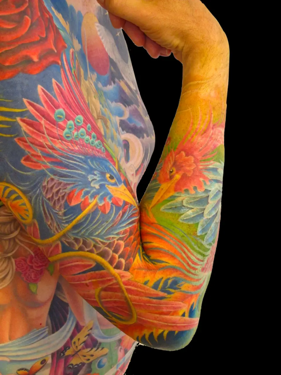 Tatuaje a color estilo manga de dos fénix besándose, en el brazo