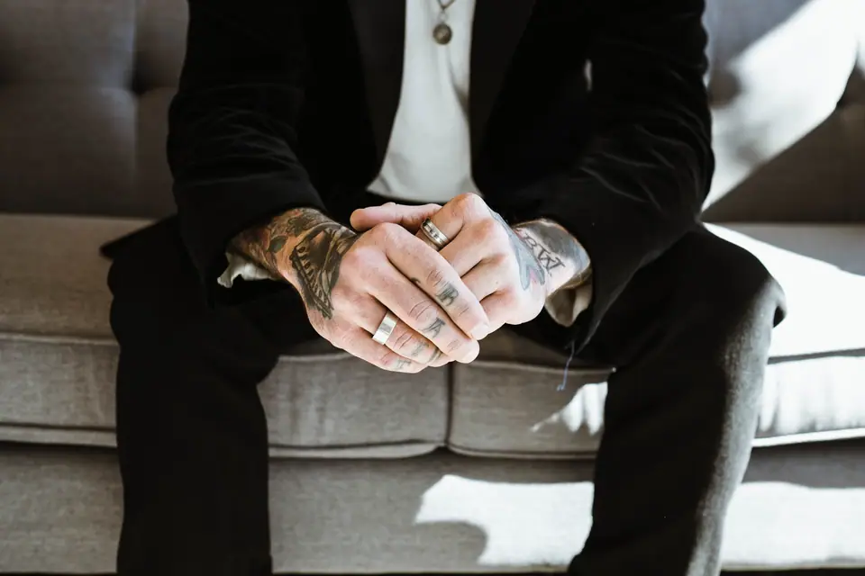 Manos de un hombre con tatuajes en varios dedos, entrelazadas sobre las piernas